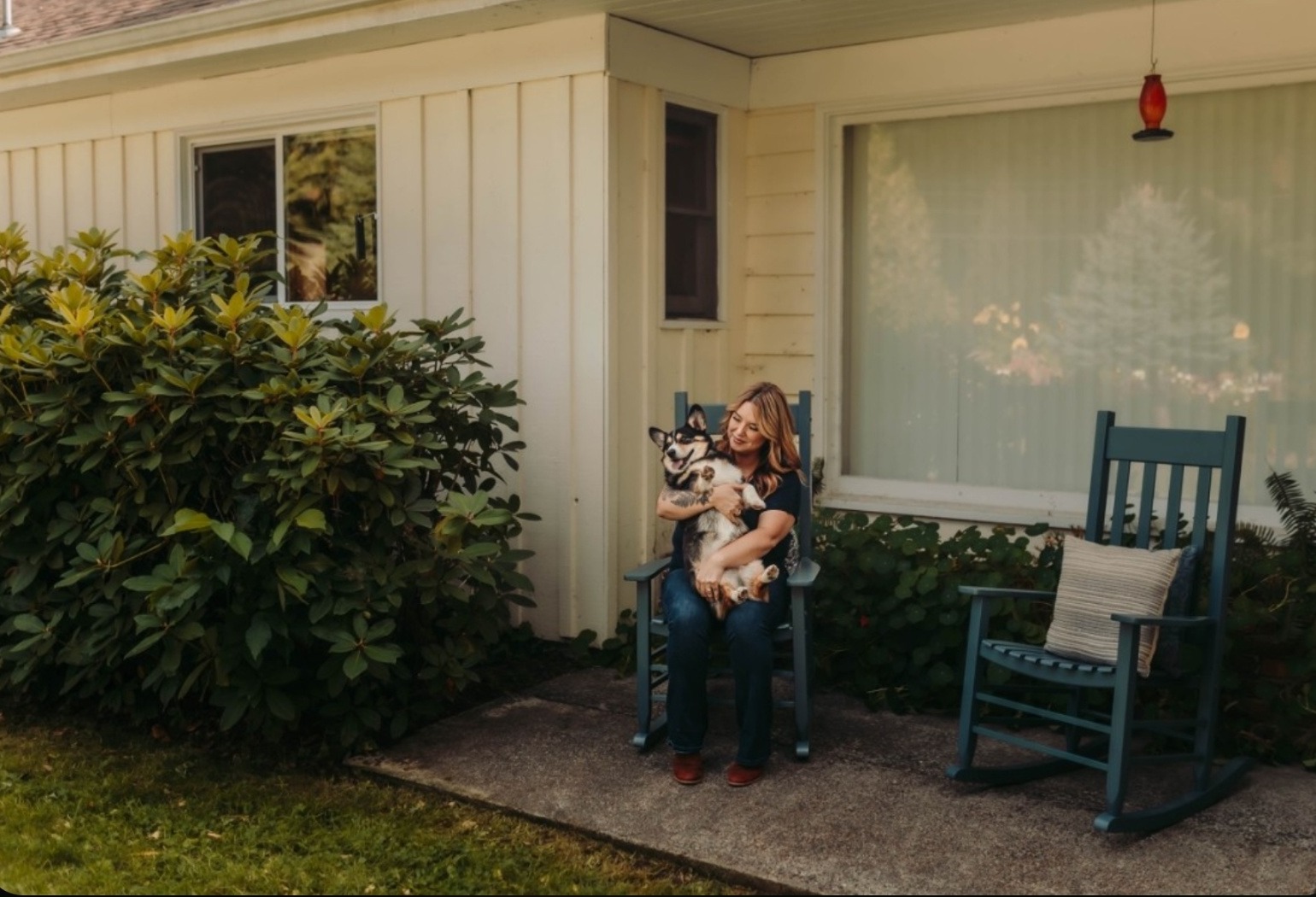 Rachel sitting in a blue rocking chair on her farmhouse porch, holding her corgi in her lap
